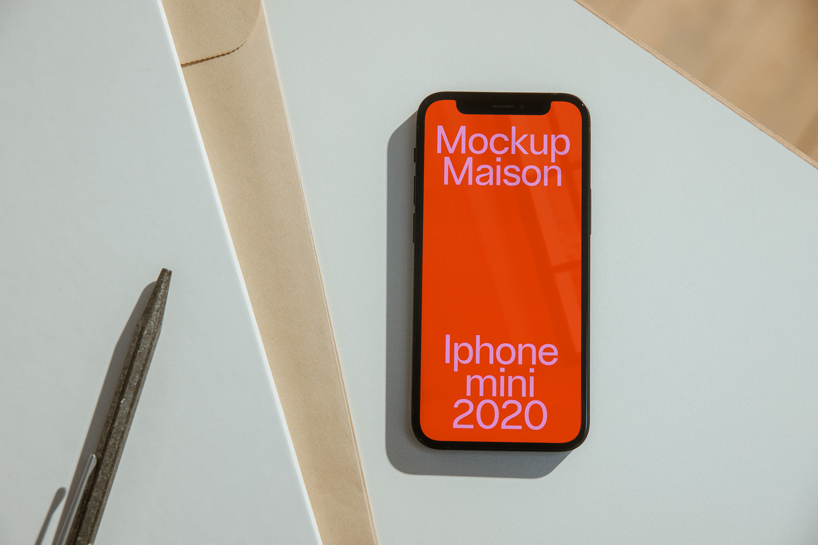 PSD mockup of an iPhone Mini 2020 with a red screen displaying "Mockup Maison" on a light background with a pen and envelope nearby.