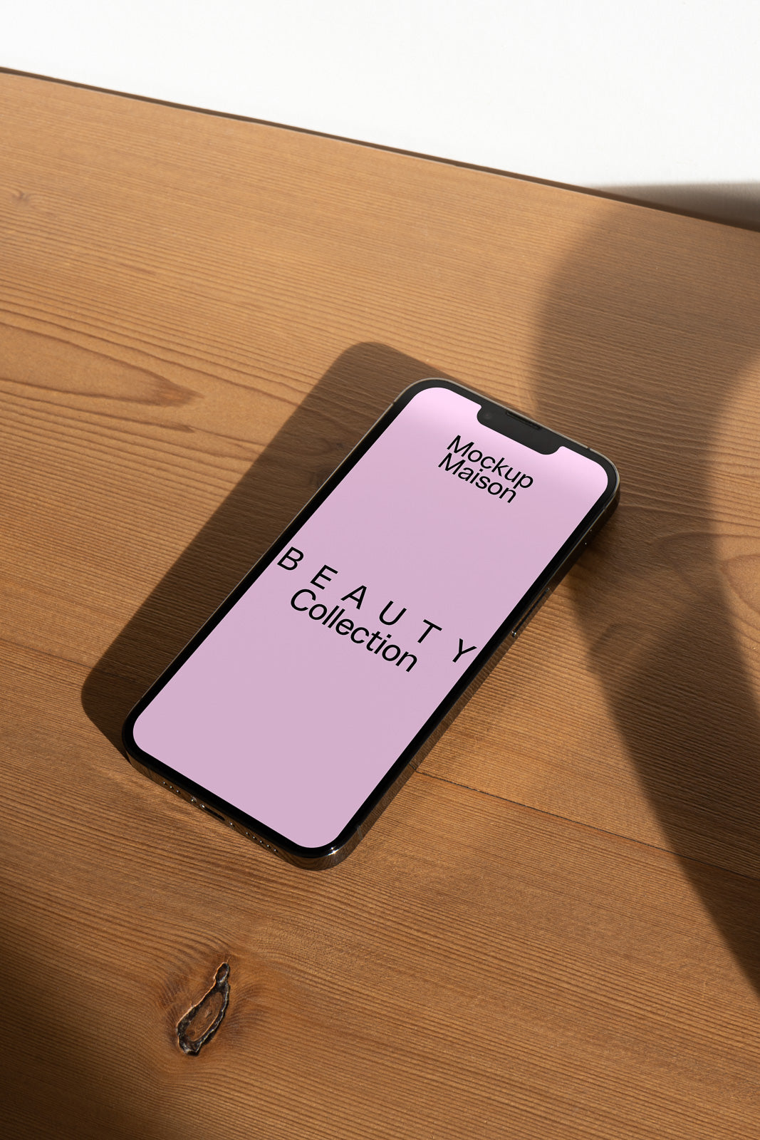 PSD mockup of a smartphone on a wooden surface displaying a pink screen with the text "Mockup Maison" and "Beauty Collection" in black.