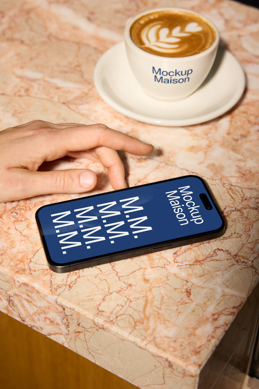 PSD mockup of a hand reaching for a smartphone with a blue screen displaying the text "Mockup Maison" on a marble countertop next to a cappuccino.