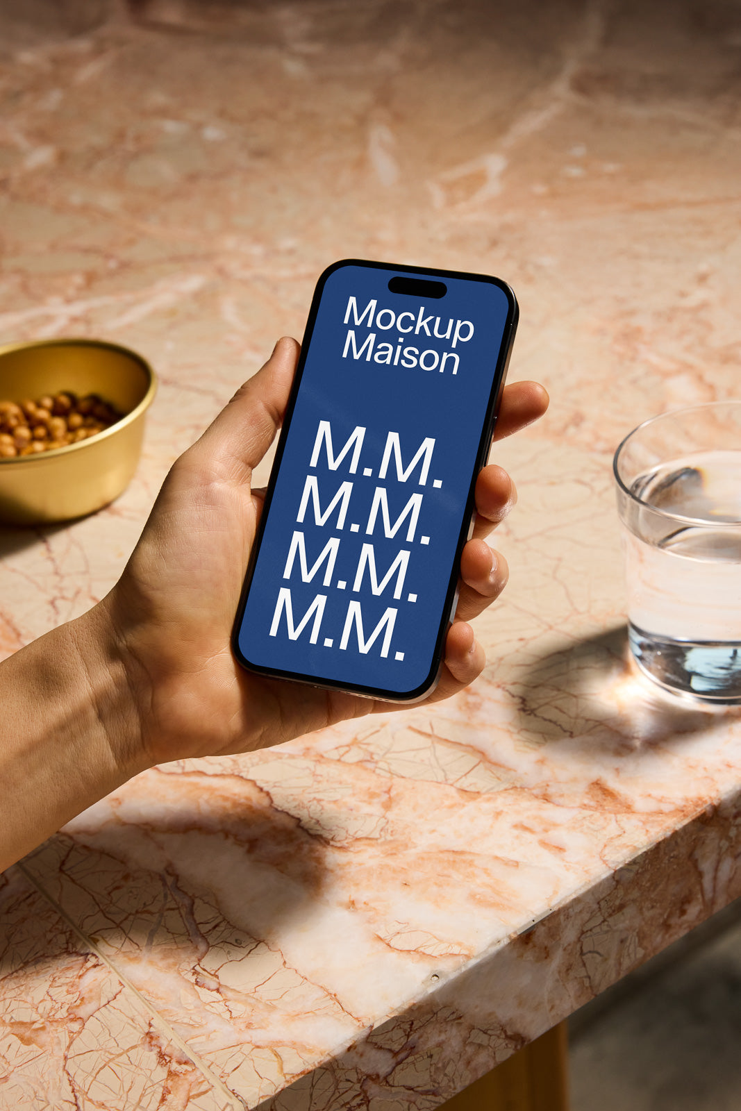 PSD mockup of a smartphone held in a hand above a marble table, displaying the text "Mockup Maison" on the screen. A glass and bowl are nearby.