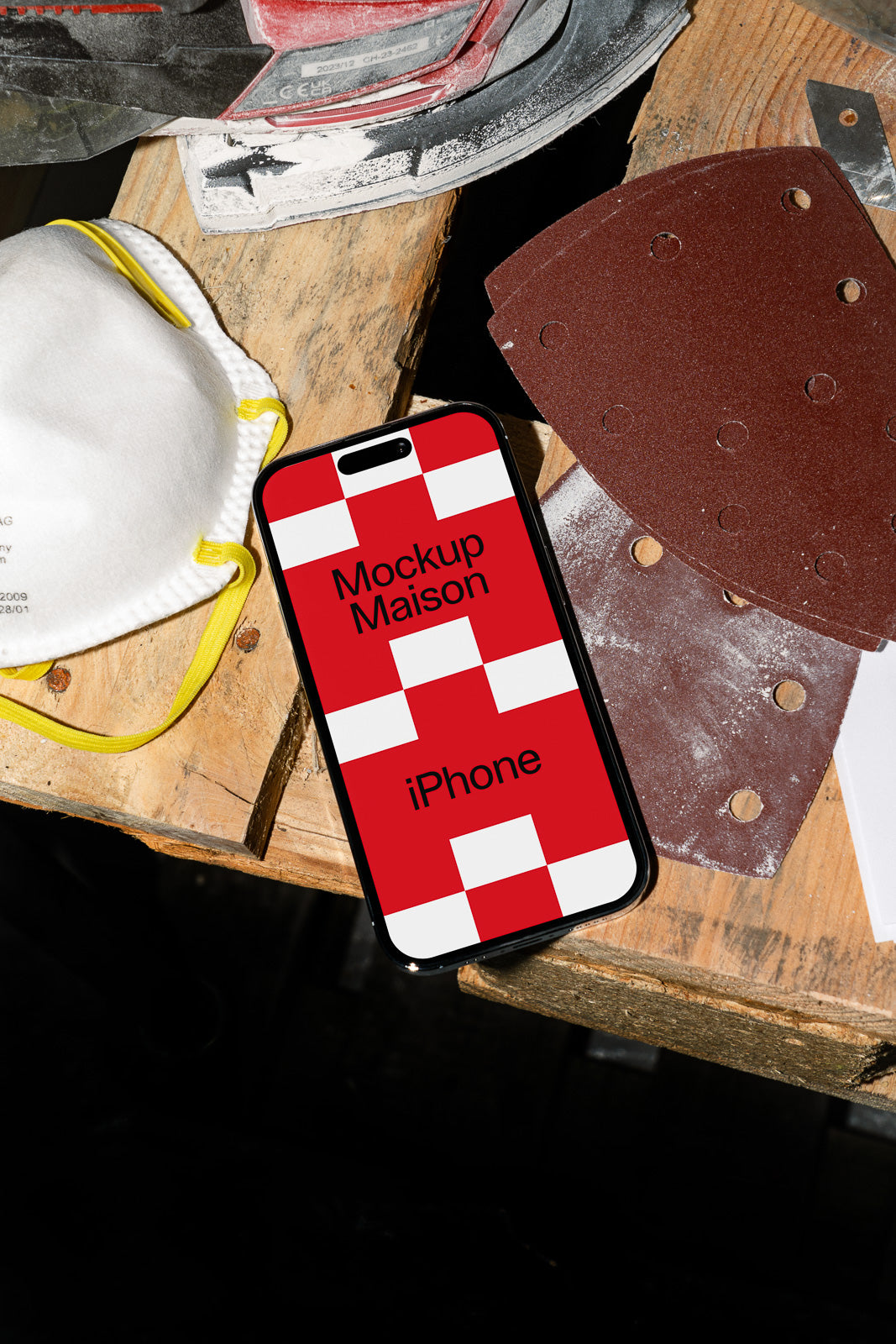 PSD mockup of an iPhone on a wooden workbench with sanding sheets and a protective mask nearby.