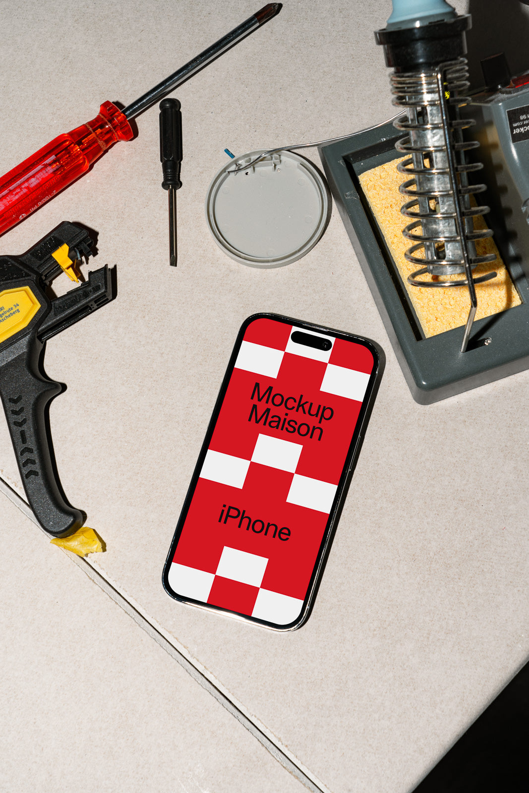 PSD mockup of an iPhone on a table surrounded by tools, featuring a checkered red and white screen displaying "Mockup Maison" text.