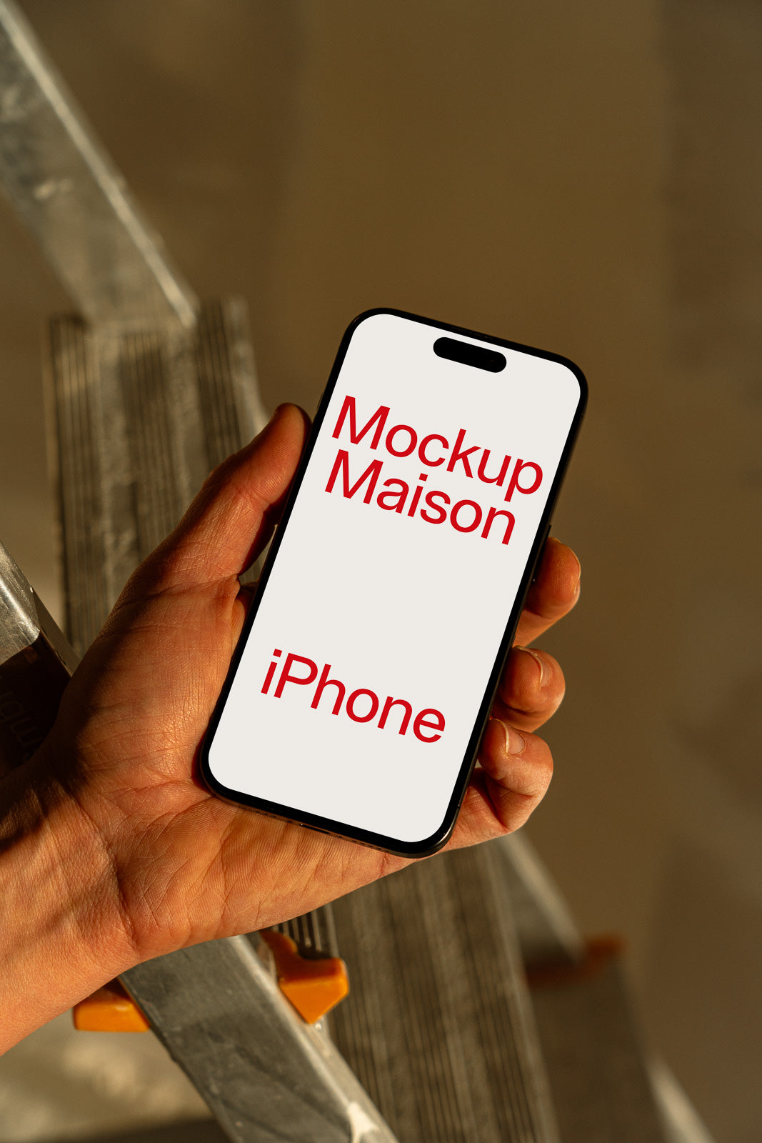 PSD mockup of hand holding a smartphone displaying the words "Mockup Maison iPhone" in red text, with a blurred background of a ladder.