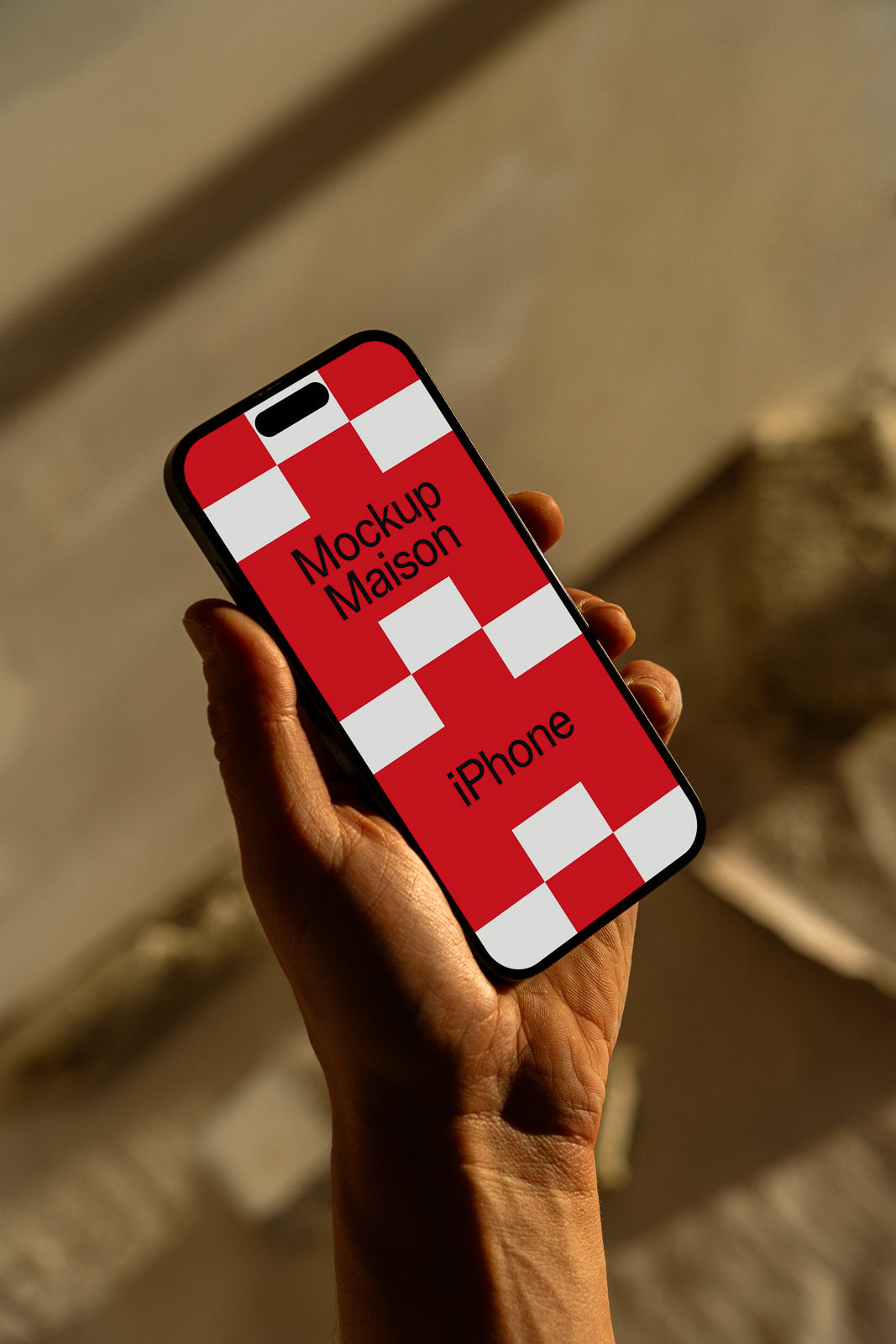 PSD mockup of a hand holding an iPhone displaying a red and white checkered screen with the text "Mockup Maison iPhone" in a well-lit environment.