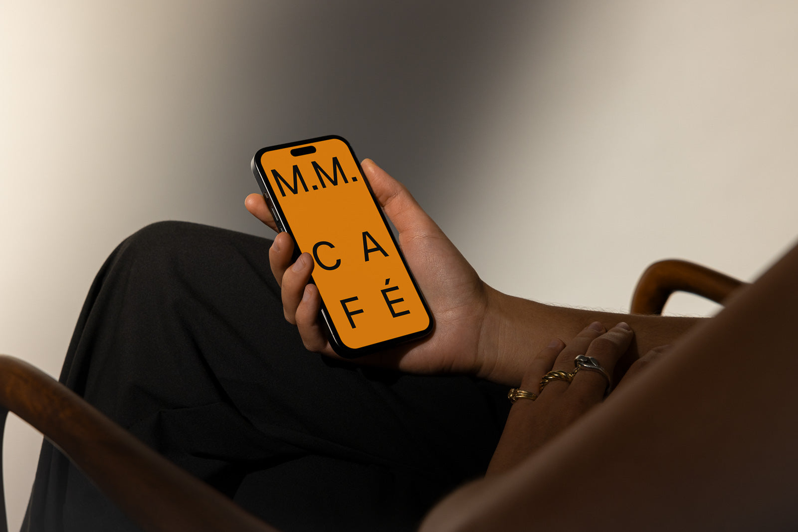 PSD mockup of a smartphone held by a person sitting in an armchair, displaying an orange screen with text "M.M. CAFÉ."