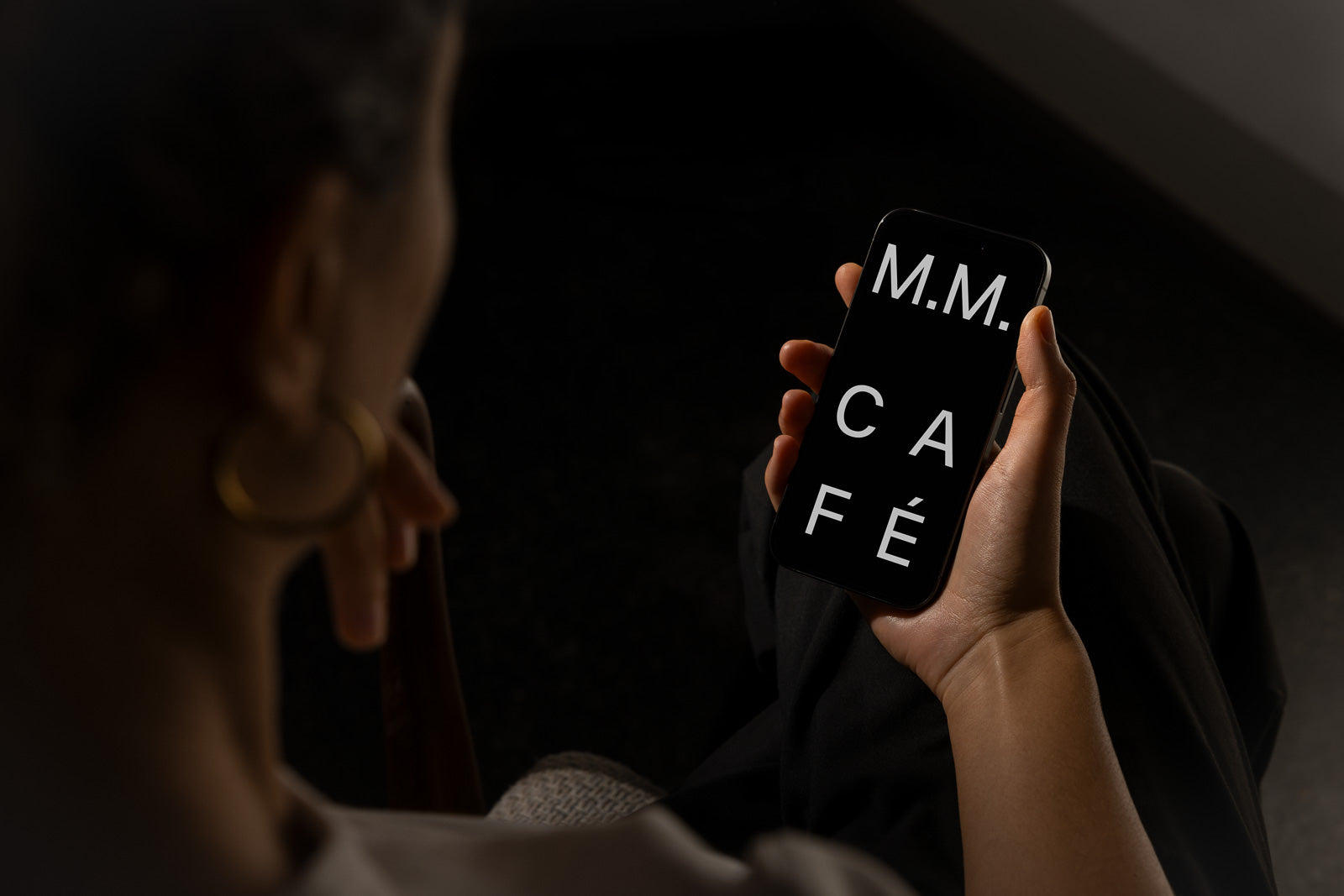 PSD mockup of a person holding a smartphone displaying the text "M.M. CAFÉ" on a dark screen. The background is softly blurred, creating a focused view on the device.