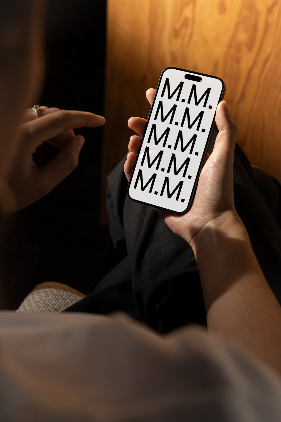 PSD mockup of a person holding a smartphone displaying a repeating pattern of the letter "M" on the screen, sitting in a dimly lit wooden setting.