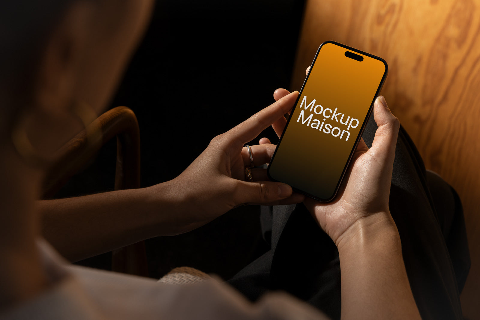 PSD mockup of a person holding a smartphone displaying “Mockup Maison” on the screen, set against a warm, wooden background in a dimly lit space.