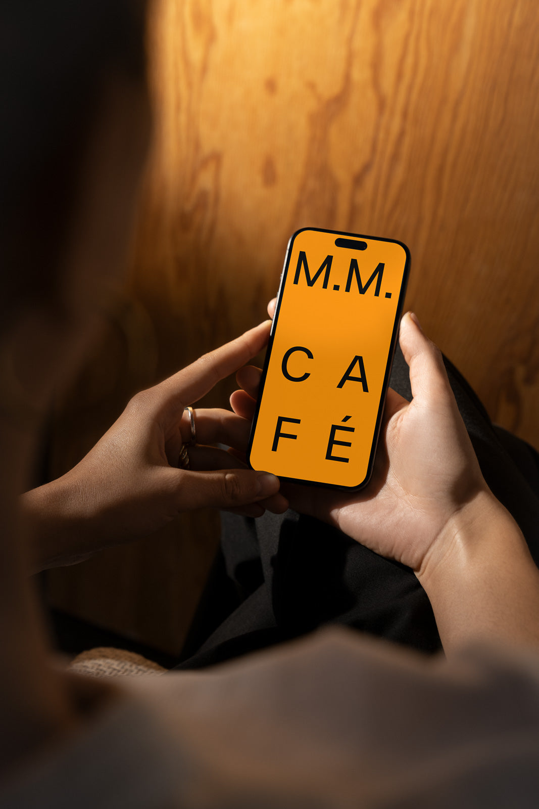 PSD mockup of a person holding a smartphone displaying the text "M.M. Café" in large, black font on an orange background.