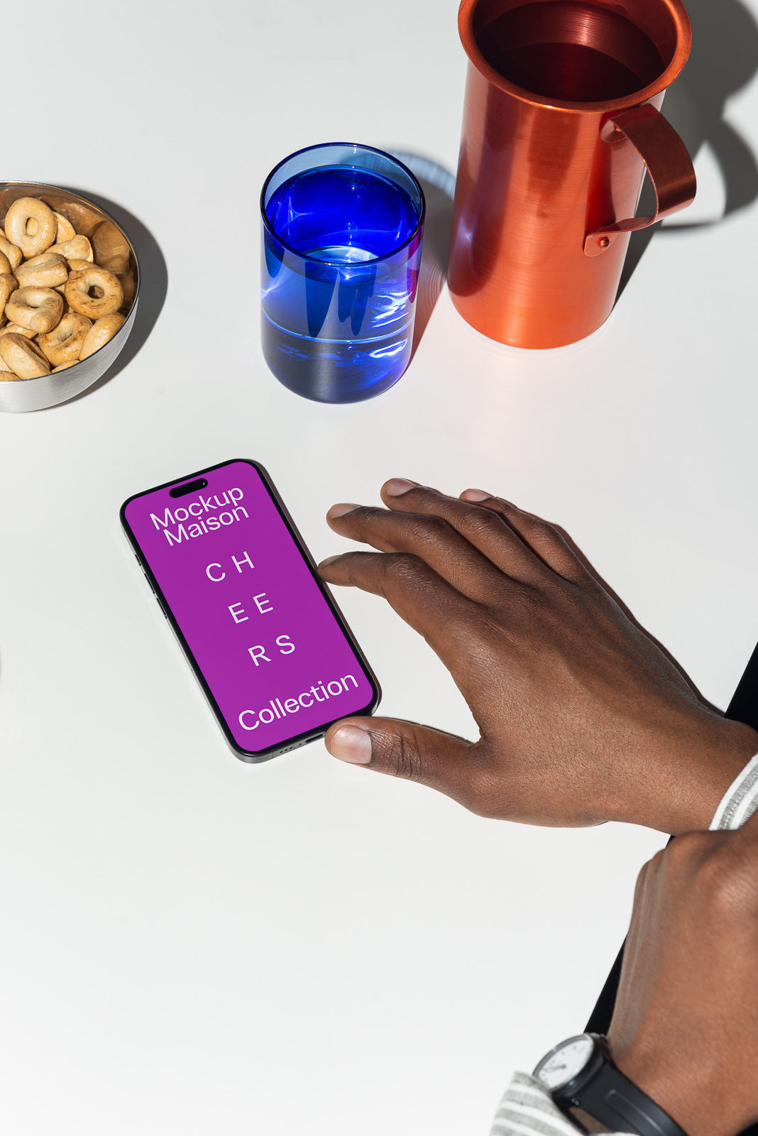 PSD mockup of a smartphone displaying "Mockup Maison Cheers Collection" on a vibrant purple screen, surrounded by a hand, snacks, blue glass, and orange jug on a white surface.