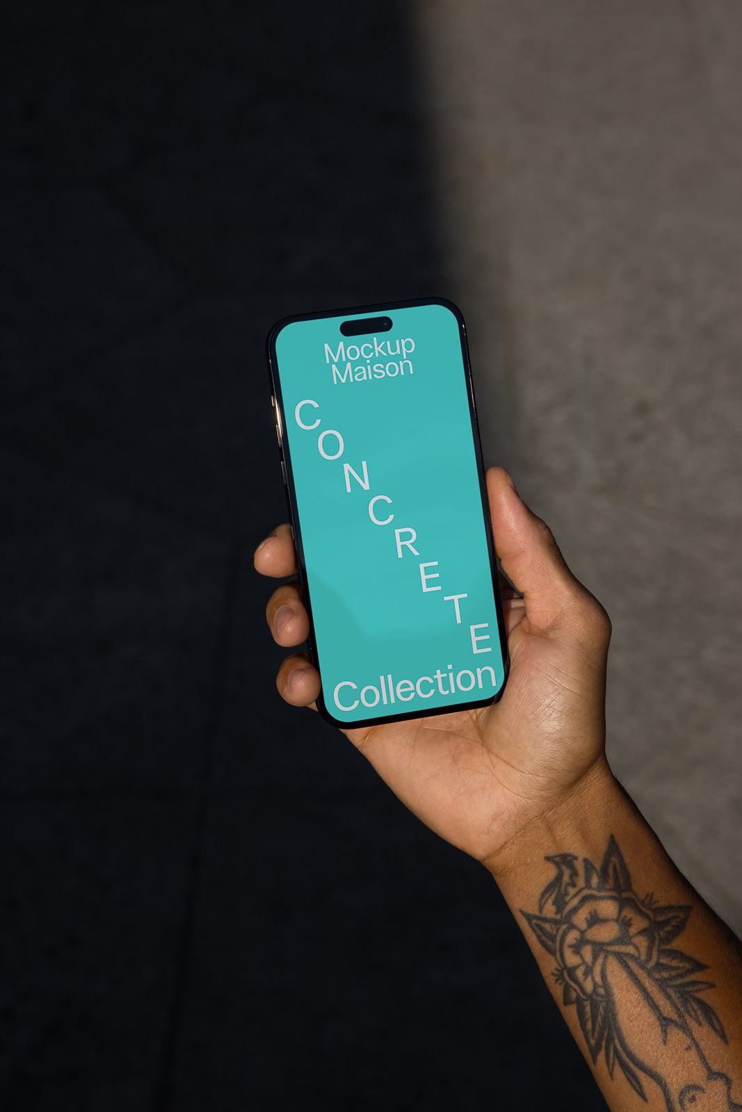PSD mockup of a smartphone held in a tattooed hand displaying a blue screen with text "Mockup Maison Concrete Collection" on a textured background.