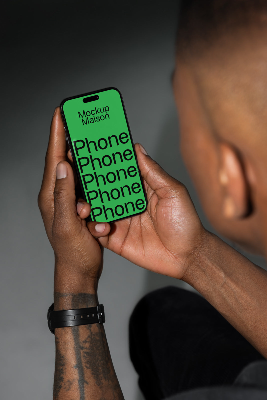 PSD mockup of a person holding a smartphone displaying a green screen with repeated bold black text reading "Phone." The screen also shows the words "Mockup Maison." The person has a visible tattoo and wears a black wristwatch.