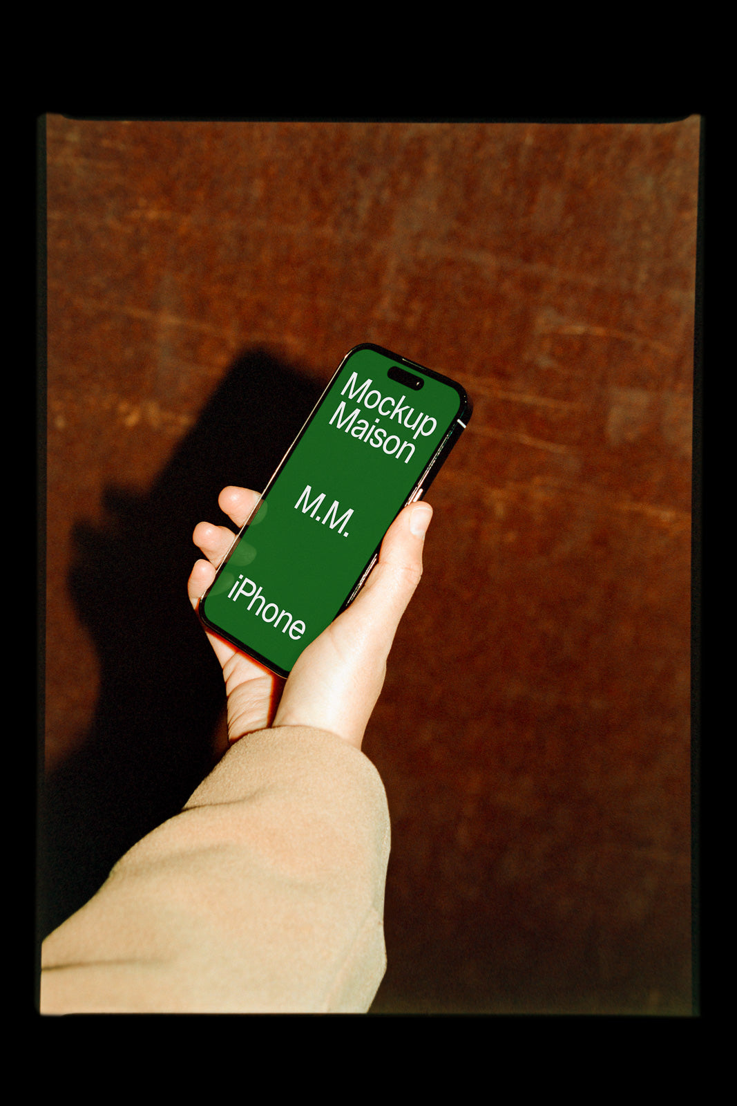 PSD mockup of a hand holding an iPhone against a textured wall, displaying a green screen with text "Mockup Maison", "M.M.", and "iPhone".