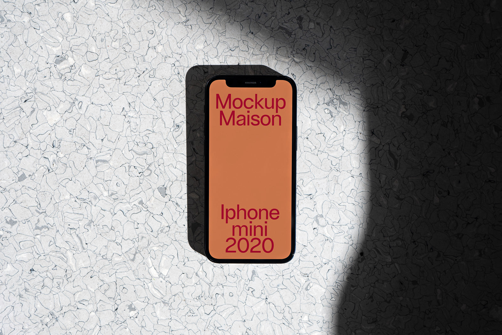 PSD mockup of an iPhone mini 2020 displaying "Mockup Maison" on an orange screen, set against a textured concrete surface with light and shadow play.