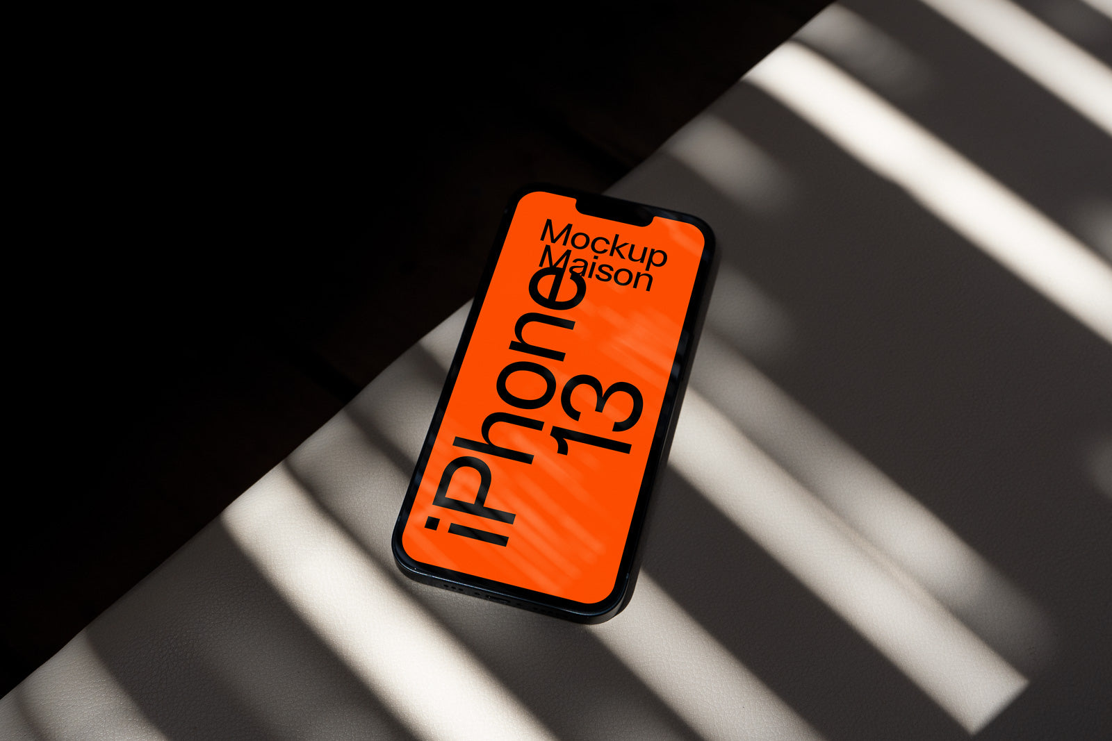 PSD mockup of an iPhone 13 on a surface with shadow patterns, displaying "Mockup Maison" on a bright orange screen.