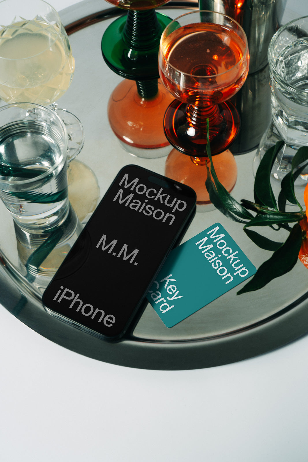 PSD mockup of a smartphone and key card on a mirrored tray with colorful drink glasses and green leaves nearby.
