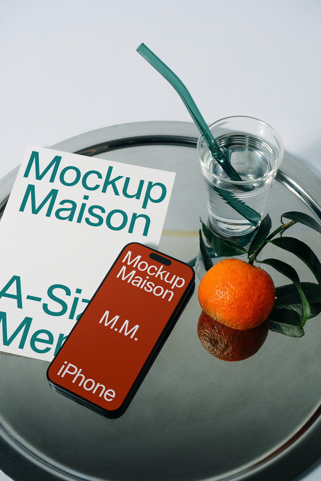 PSD mockup of an iPhone with "Mockup Maison" text next to a flyer, glass with a green straw, and an orange on a silver tray.