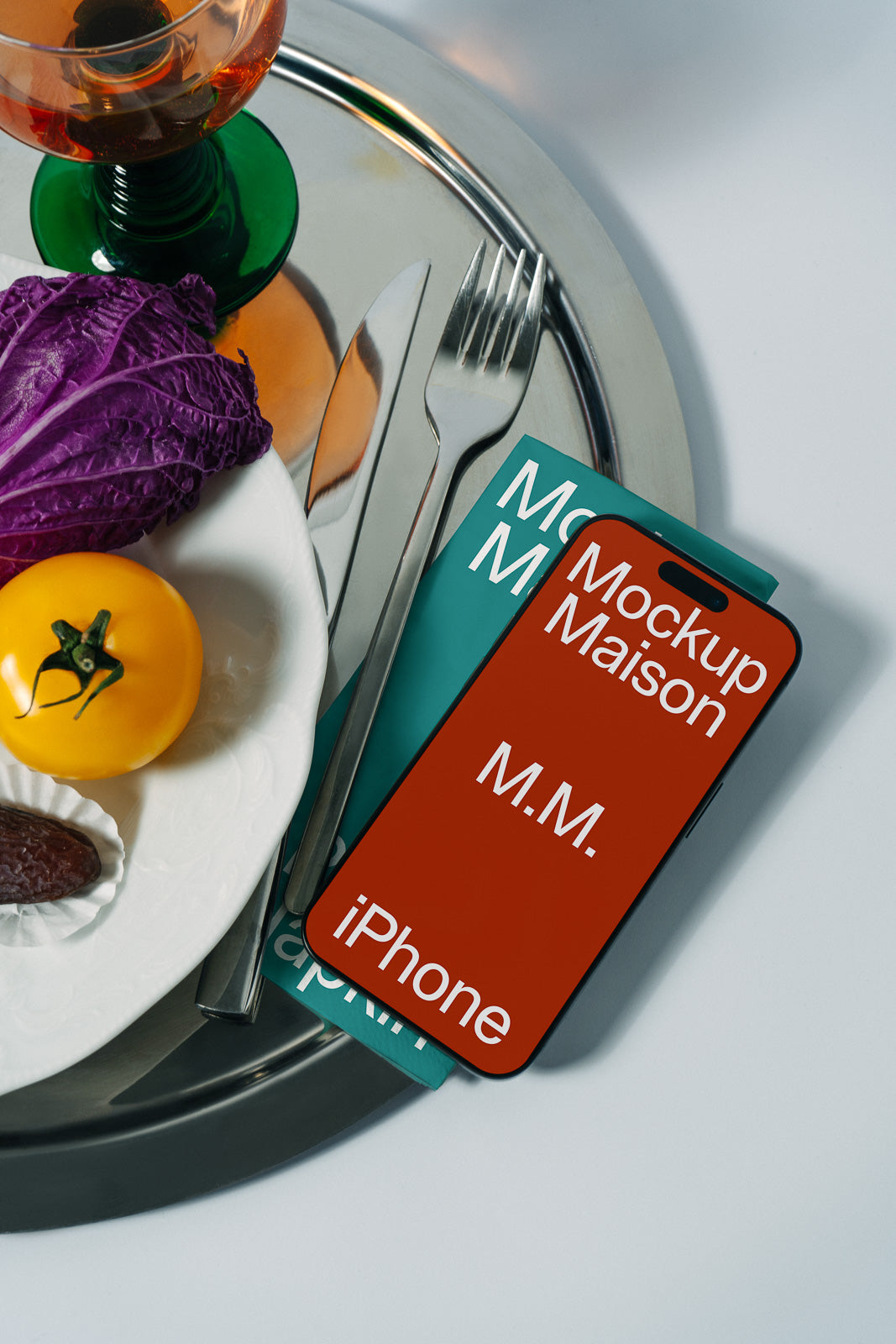 PSD mockup of a smartphone with a red screen displaying text "Mockup Maison" and "iPhone" placed on a silver tray next to a fork, knife, cabbage, and yellow tomato.