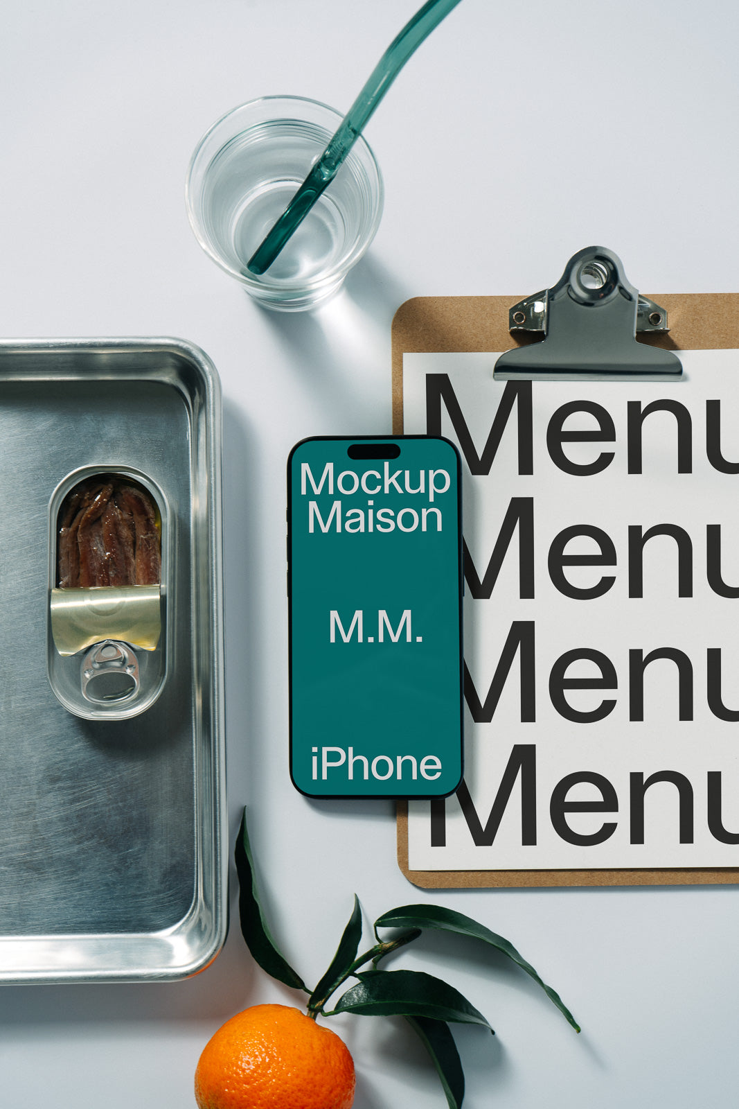 PSD mockup of an iPhone displaying "Mockup Maison" on a green screen, placed beside a clipboard with a menu, a can of food, a glass of water, and an orange.