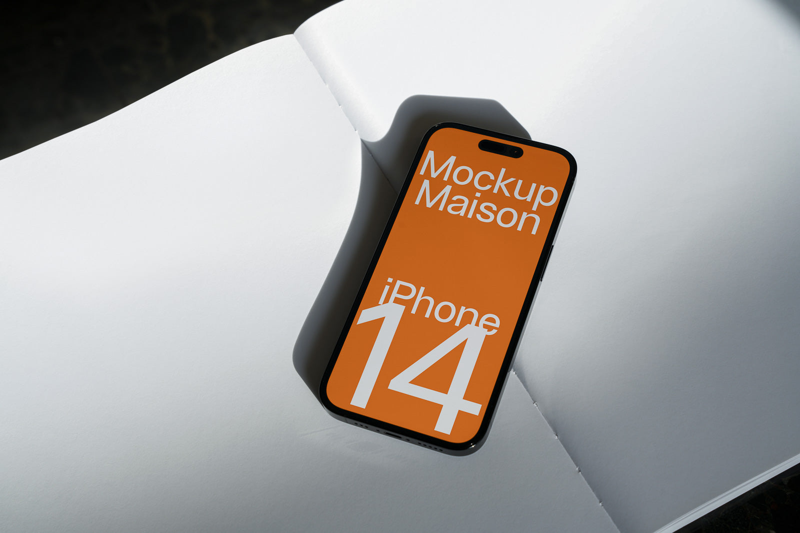 PSD mockup of an iPhone 14 displaying "Mockup Maison" on an orange screen, placed on an open white book with shadows.