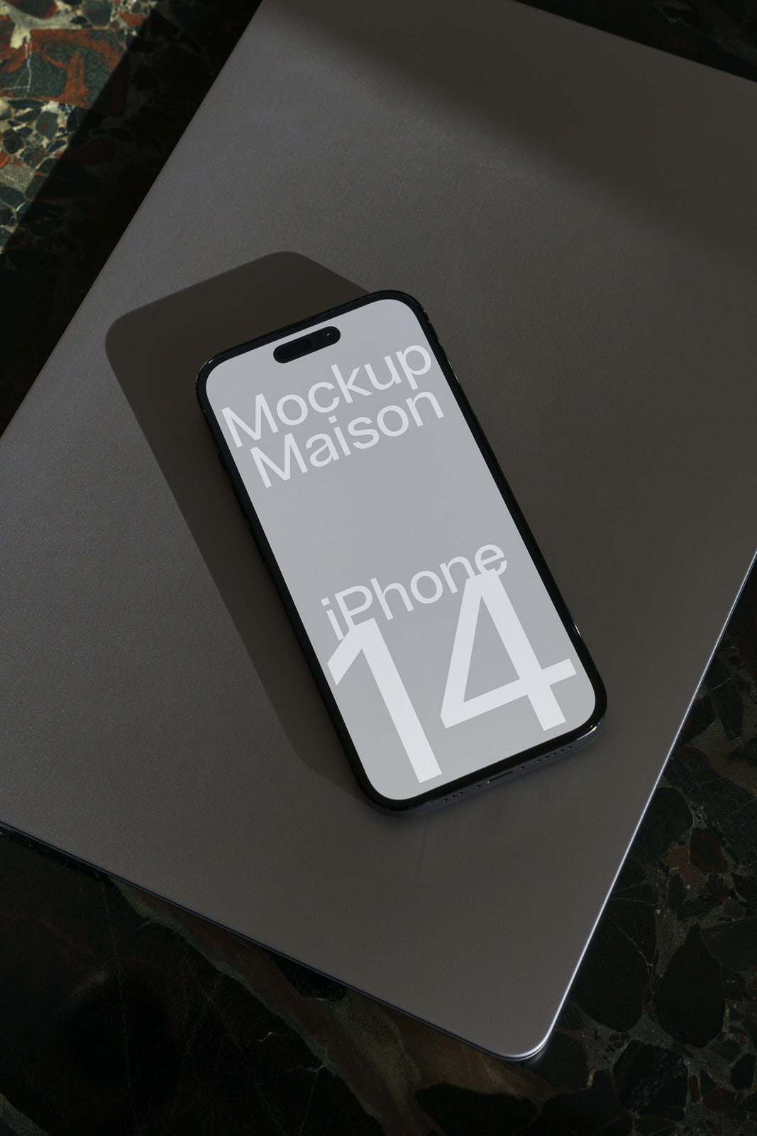 PSD mockup of an iPhone 14 lying on a closed laptop, displaying "Mockup Maison" text on the phone screen, set against a dark, patterned surface.
