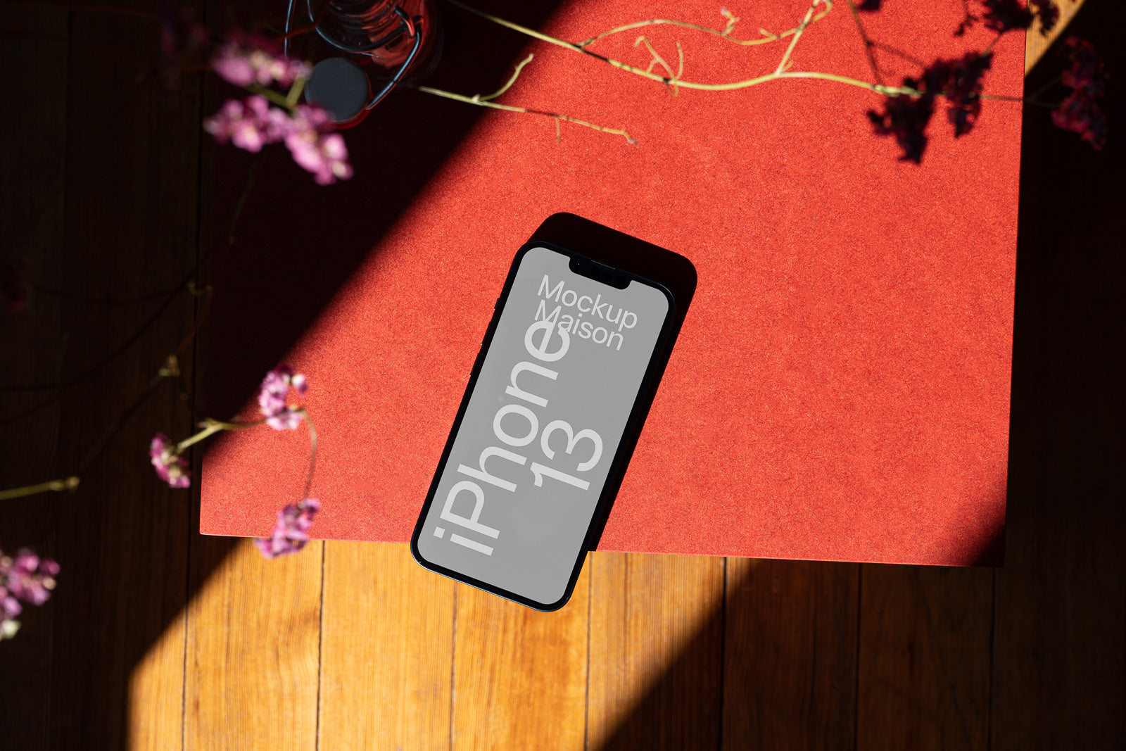 PSD mockup of an iPhone 13 on a red surface, with sunlight casting shadows and flowers blurred in the foreground.