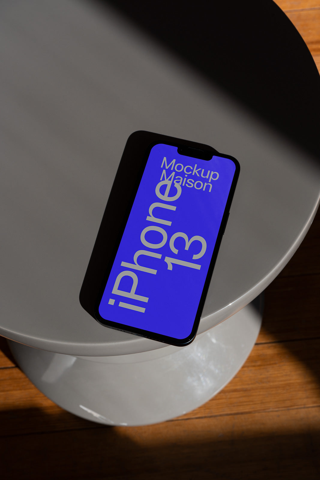 PSD mockup of an iPhone 13 on a circular gray table with blue screen displaying text "Mockup Maison" in light and shadow.