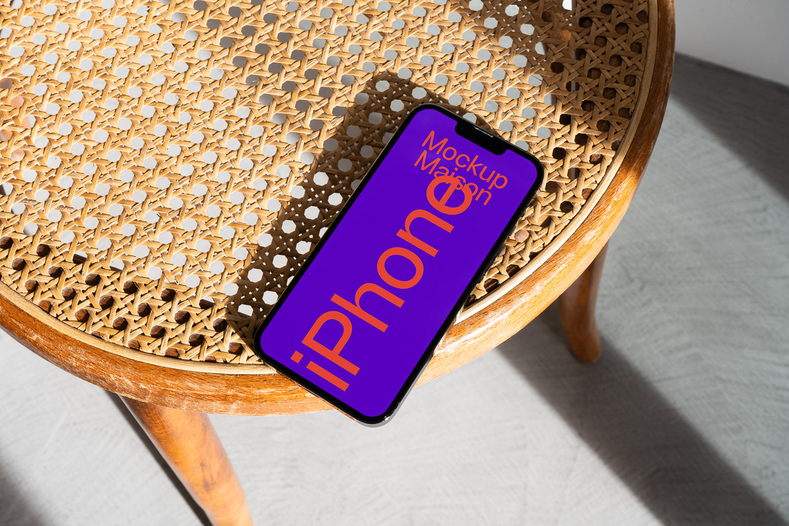PSD mockup of a smartphone with a vibrant purple screen displaying text, set on a wooden chair with woven patterns and sunlight casting shadows.