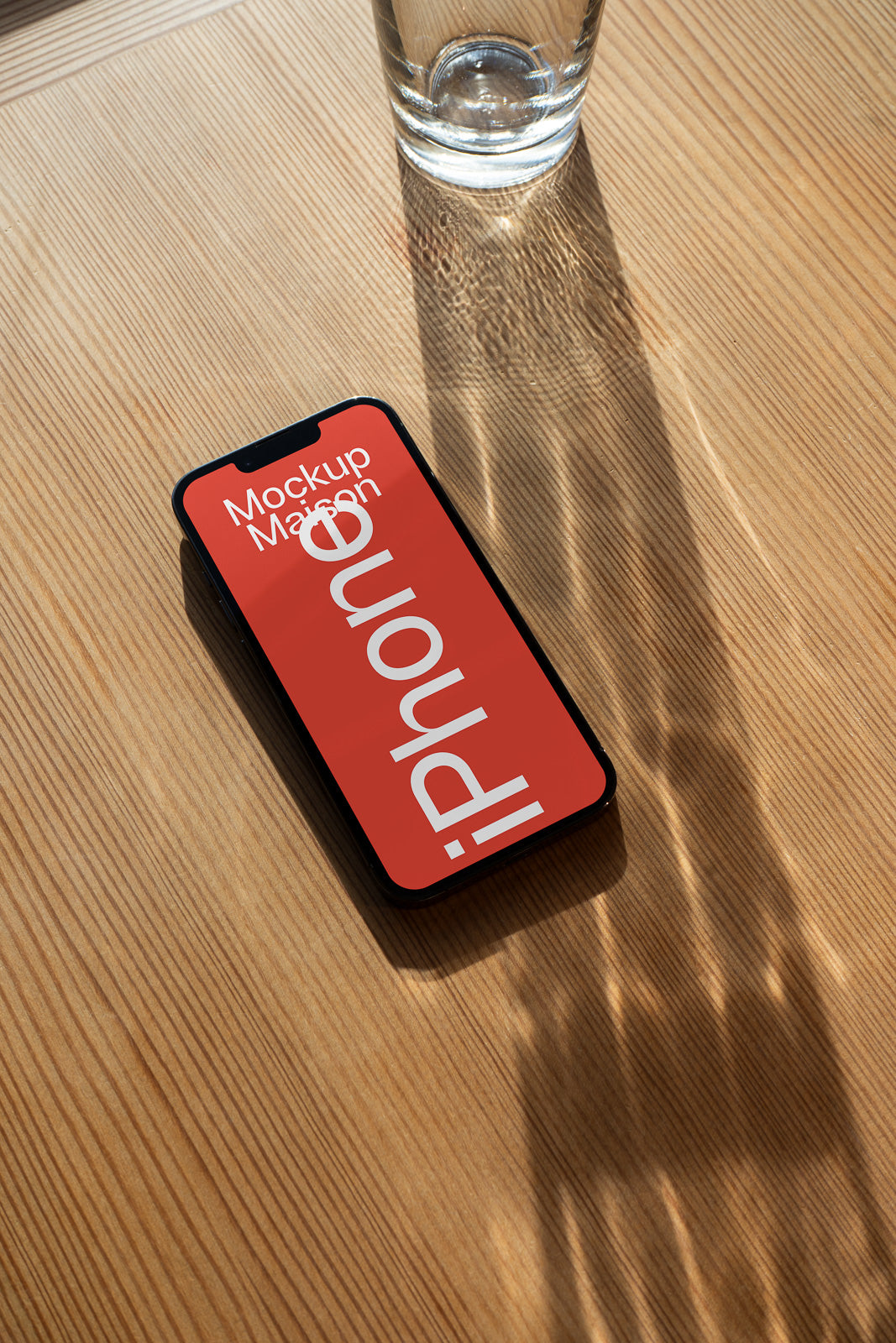 PSD mockup of a smartphone displaying a red screen with white text on a wooden table beside a glass of water reflecting sunlight.
