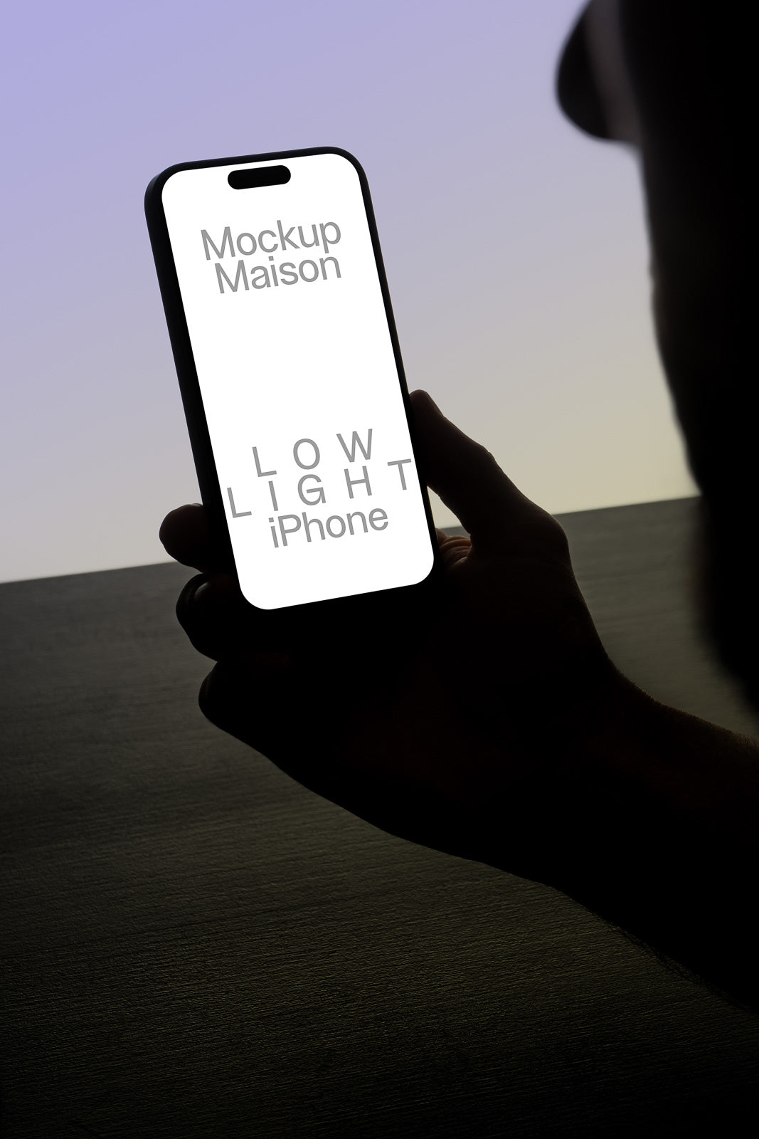 PSD mockup of a hand holding a smartphone with a blank glowing screen displaying the text "Mockup Maison" and "Low Light iPhone," set against a dimly lit background.