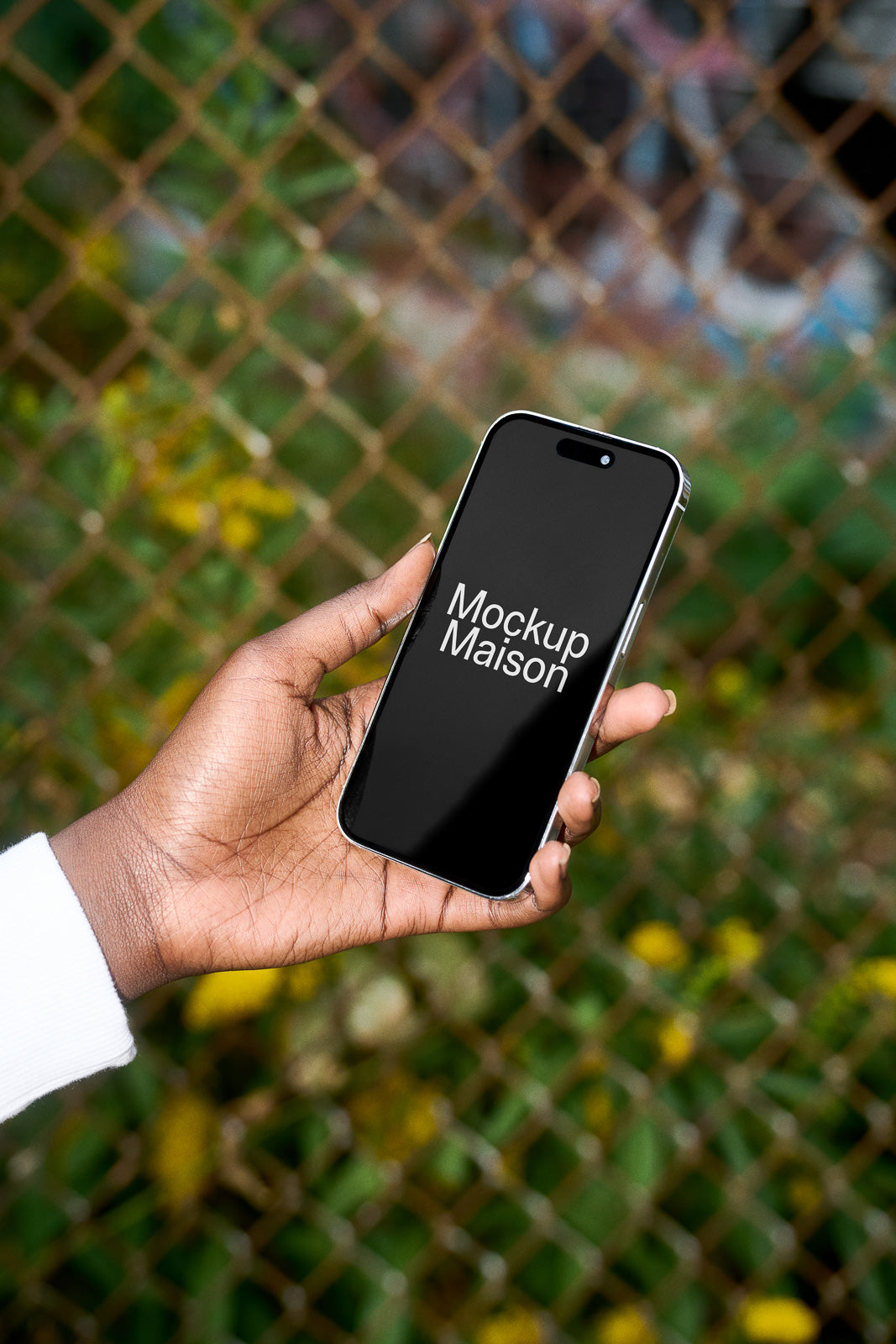 PSD mockup of a hand holding a smartphone with "Mockup Maison" on the screen, set against a blurred outdoor background.