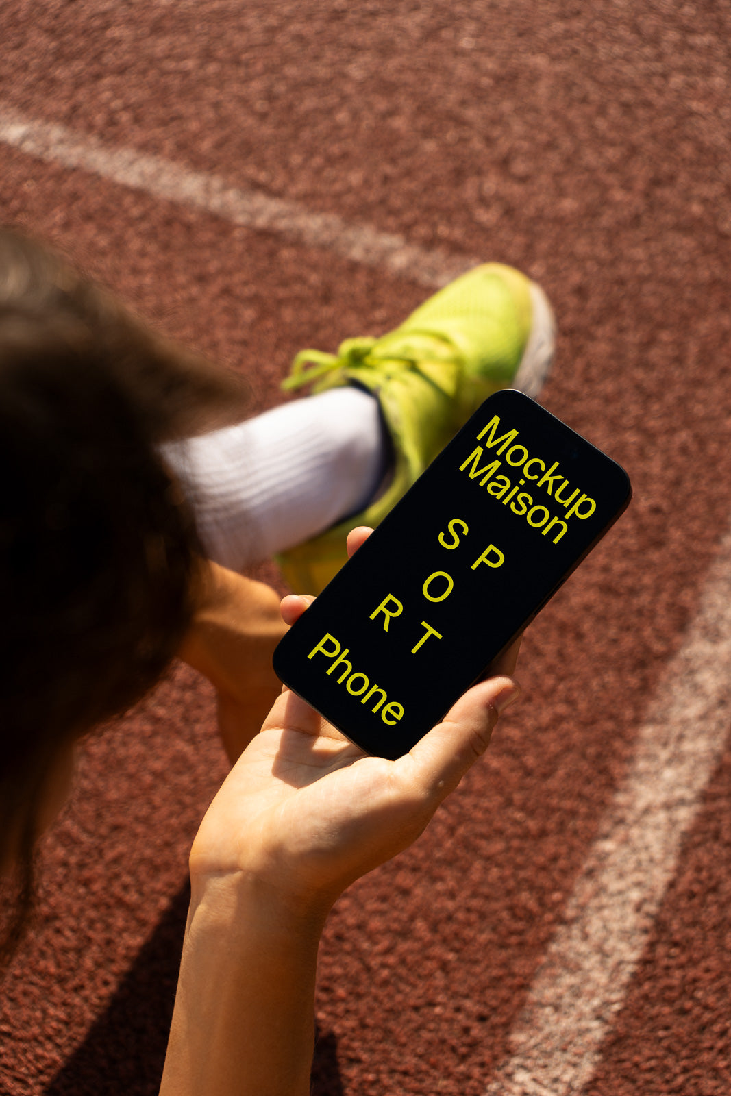 PSD mockup of a smartphone held by a person seated on a running track, displaying text in yellow on the screen.