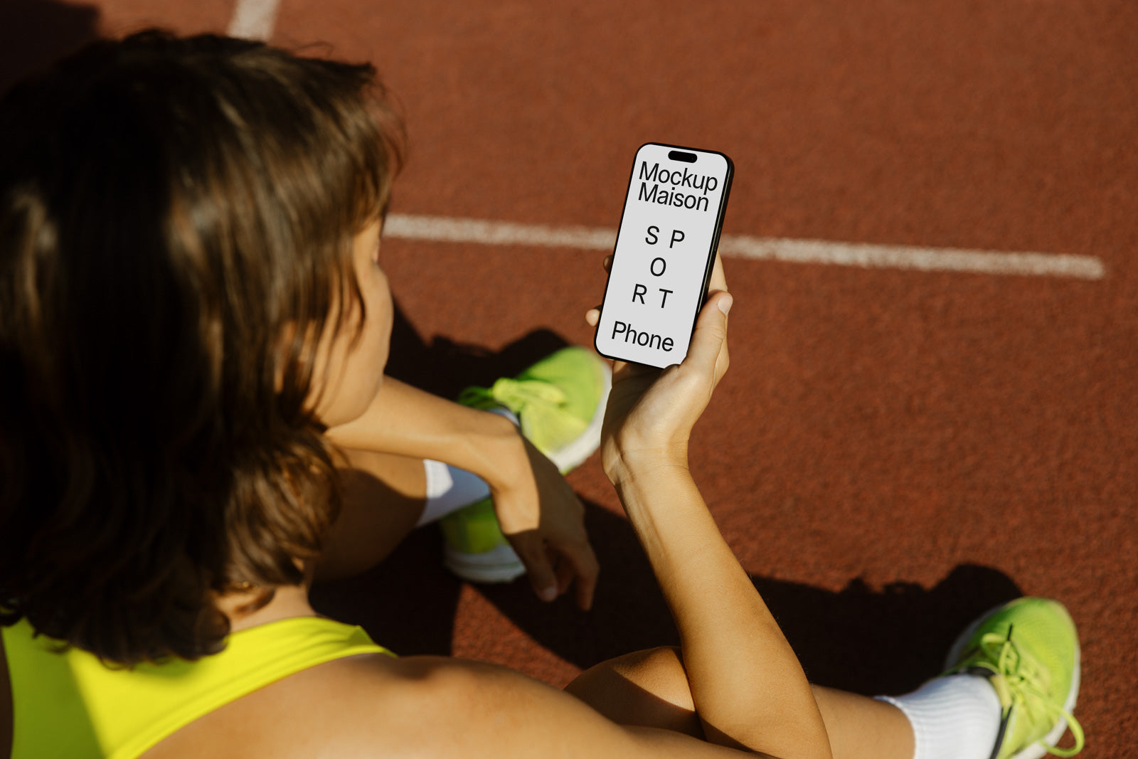 PSD mockup of a person sitting on a track, holding a smartphone displaying text "Mockup Maison SPORT Phone", wearing green shoes, in workout attire.