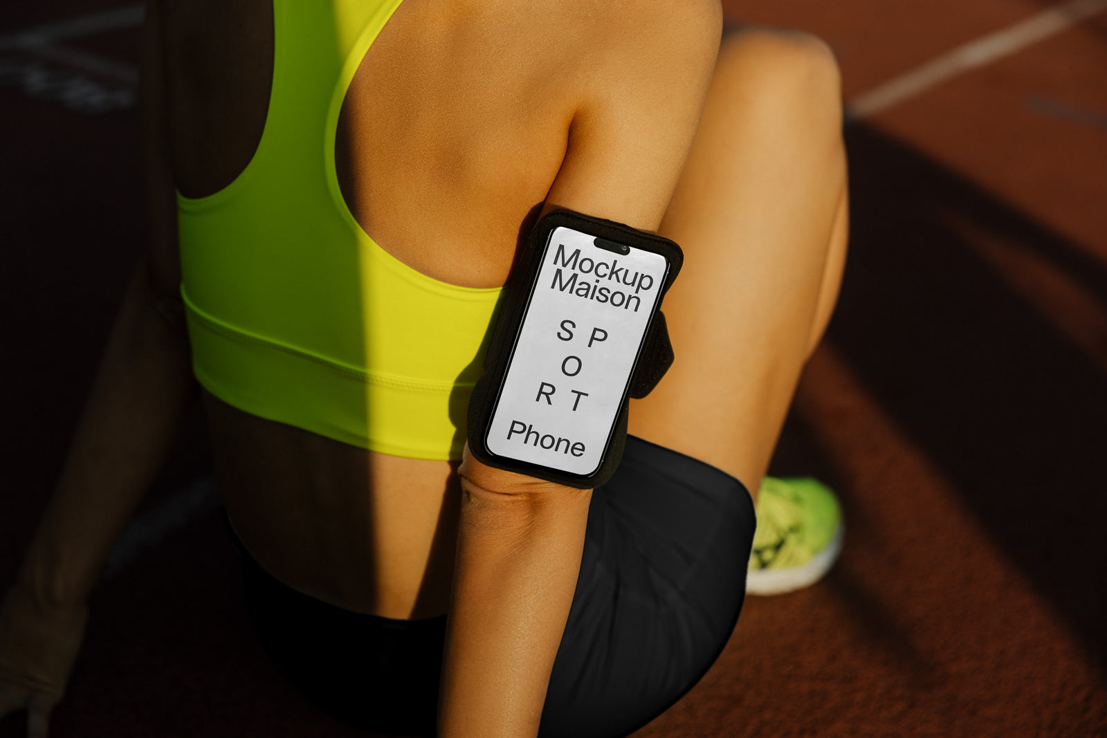 PSD mockup of a person sitting on a track wearing a yellow sports top and black armband with a phone displaying "Mockup Maison SPORT Phone."