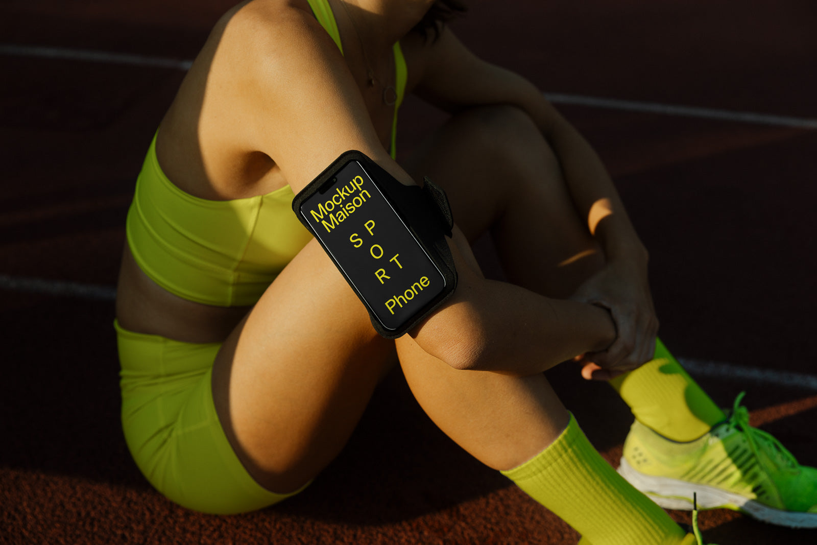 PSD mockup of a sport armband phone holder worn by an athlete in yellow workout attire, sitting on a track with sunlight cast on her.
