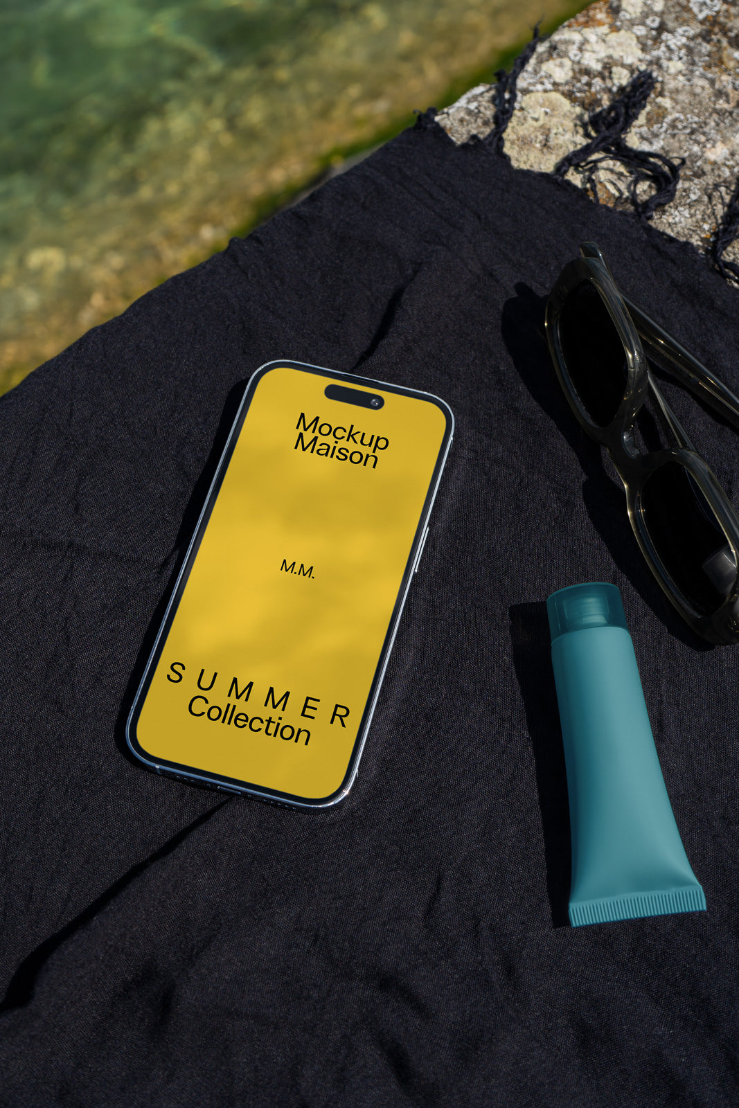 PSD mockup of a smartphone displaying "Mockup Maison" and "Summer Collection" on a yellow screen, placed on a dark cloth near sunglasses and a blue bottle.