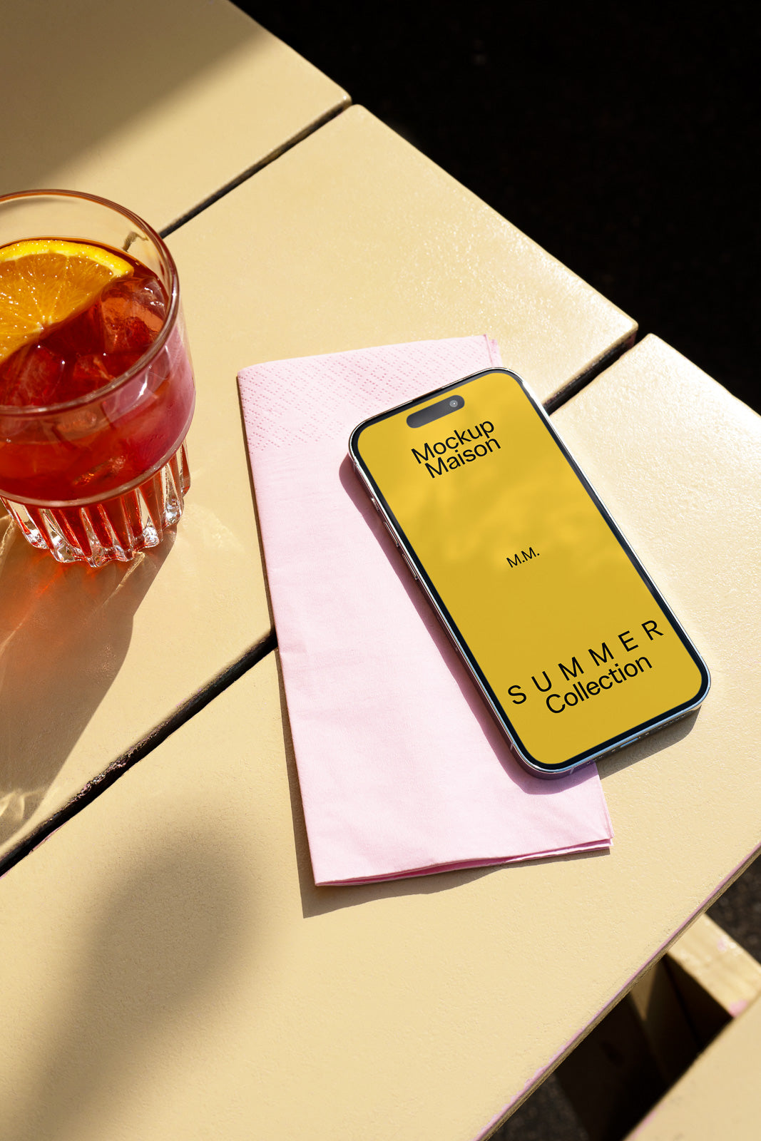 PSD mockup of a smartphone displaying "Mockup Maison Summer Collection" on a table next to a pink napkin and a juice glass with an orange slice.