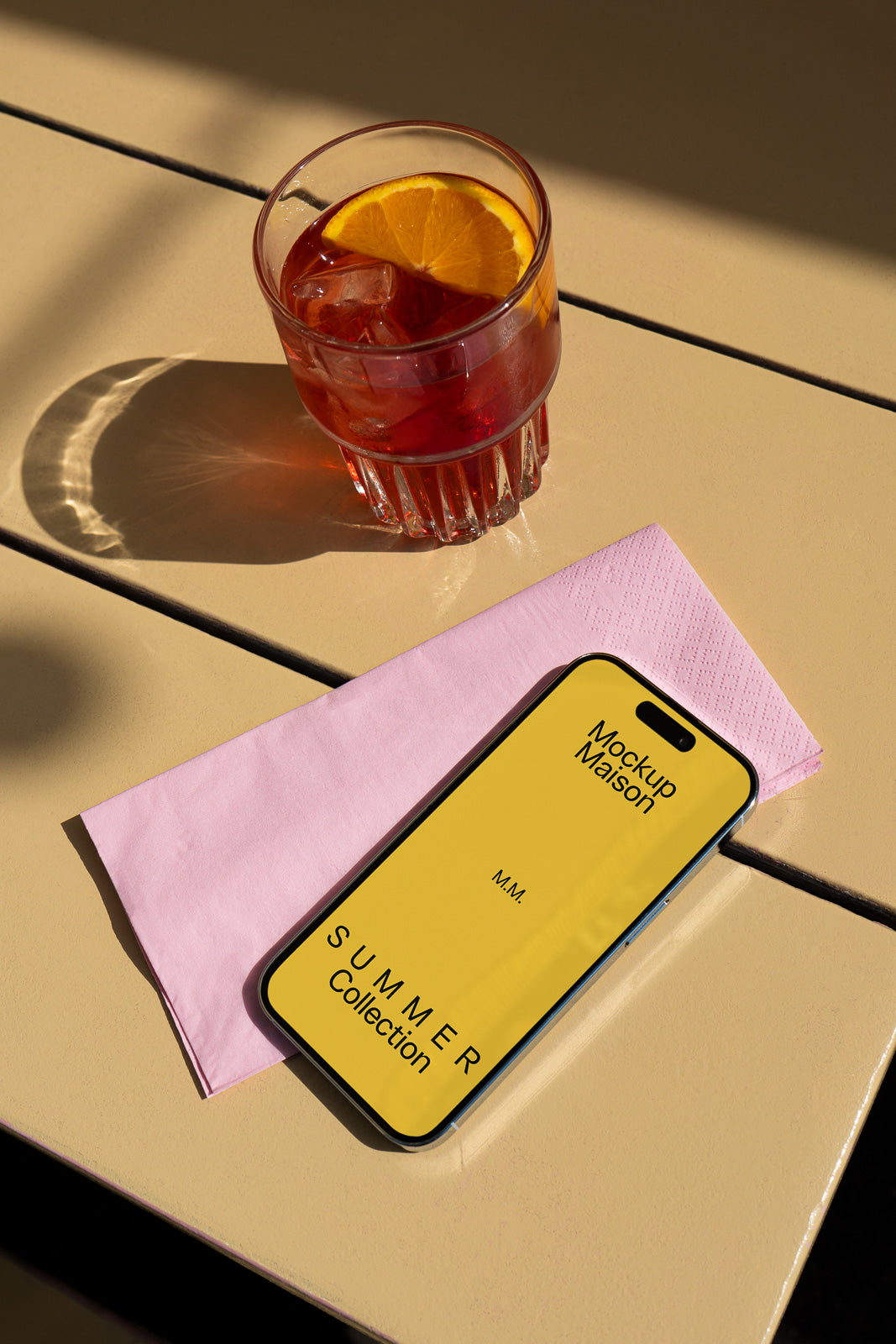 PSD mockup of a smartphone displaying a yellow screen with "Mockup Maison Summer Collection" on a table next to a pink napkin and a glass of iced drink with orange slice.