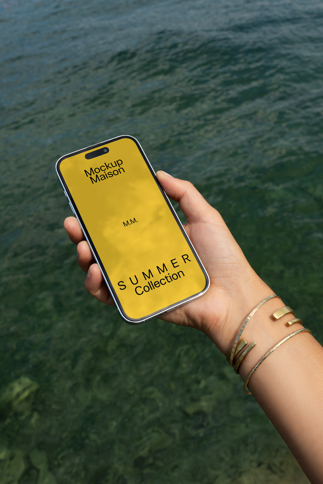 PSD mockup of a smartphone held by a hand with bracelets, displaying a yellow screen that reads "Mockup Maison Summer Collection" against a water backdrop.