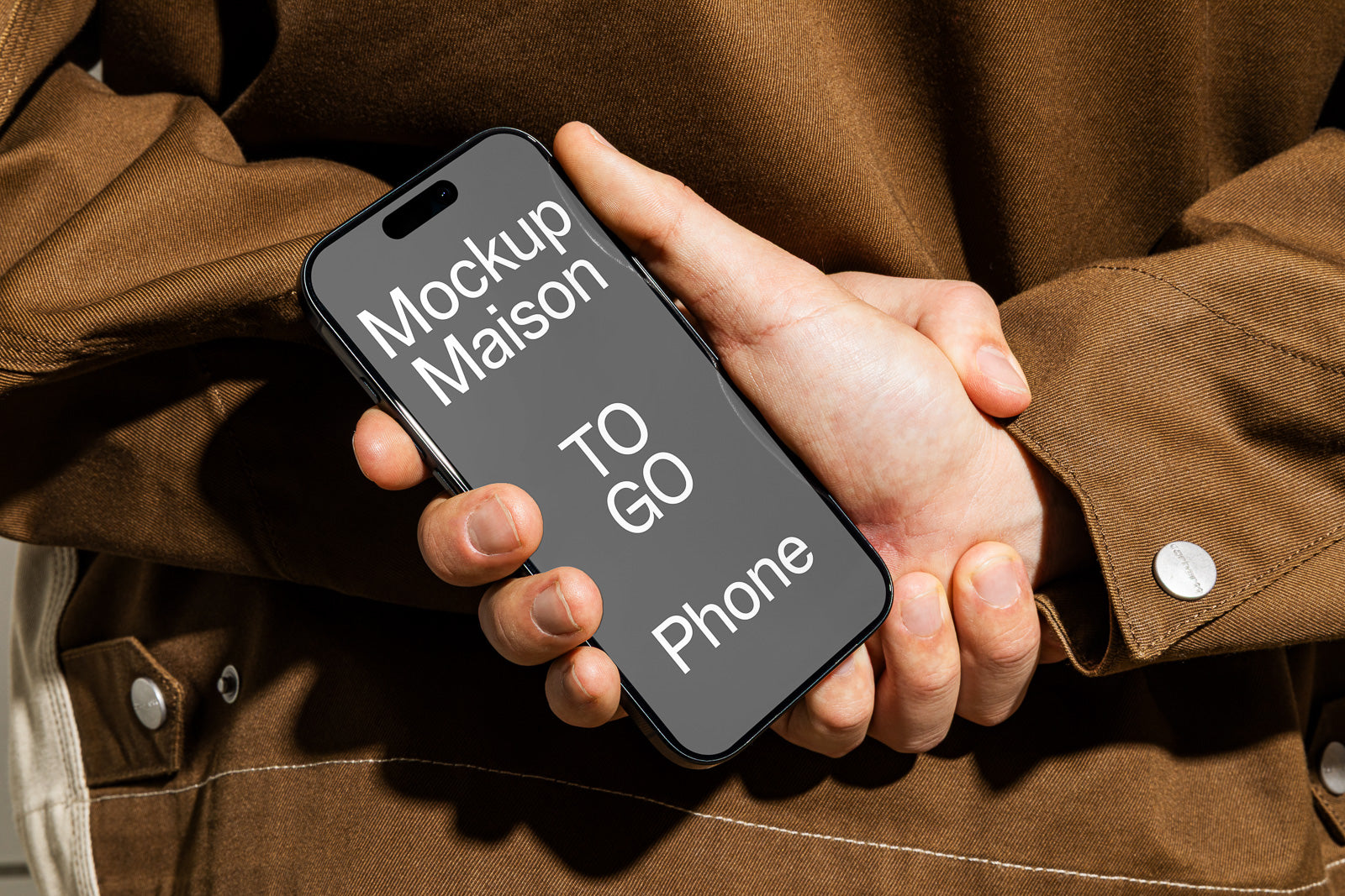 PSD mockup of a smartphone held in hands, showcasing the text "Mockup Maison TO GO Phone" on the screen against a brown fabric jacket background.