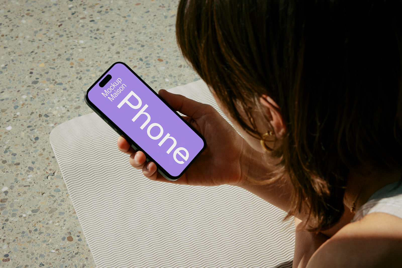 PSD mockup of a person holding a smartphone displaying a lavender screen with the word "Phone" and "Mockup Maison" while sitting on a textured surface.