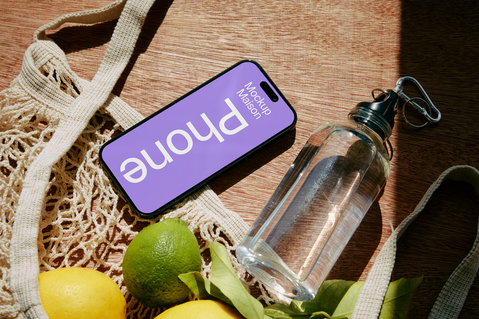 PSD mockup of a smartphone with a purple screen displaying the word "Phone," placed on a wooden surface beside a net bag with lemons, a lime, and a glass water bottle.
