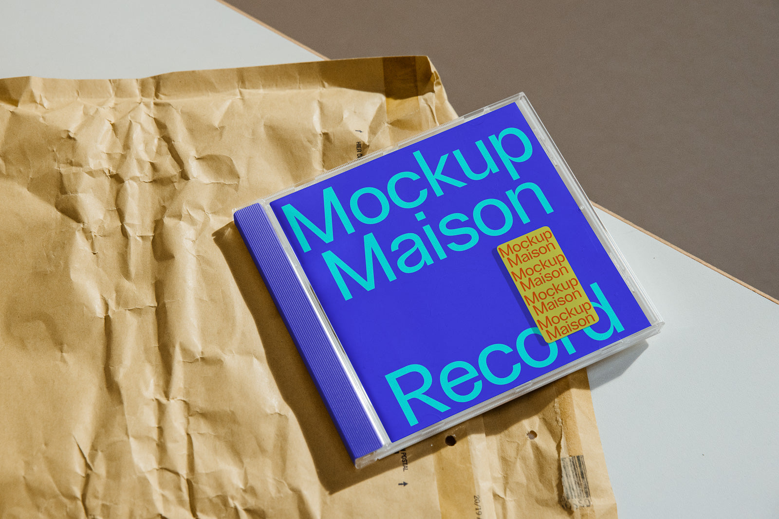 PSD mockup of a CD case labeled "Mockup Maison" in bold blue and green text, placed on crumpled brown paper on a flat surface.