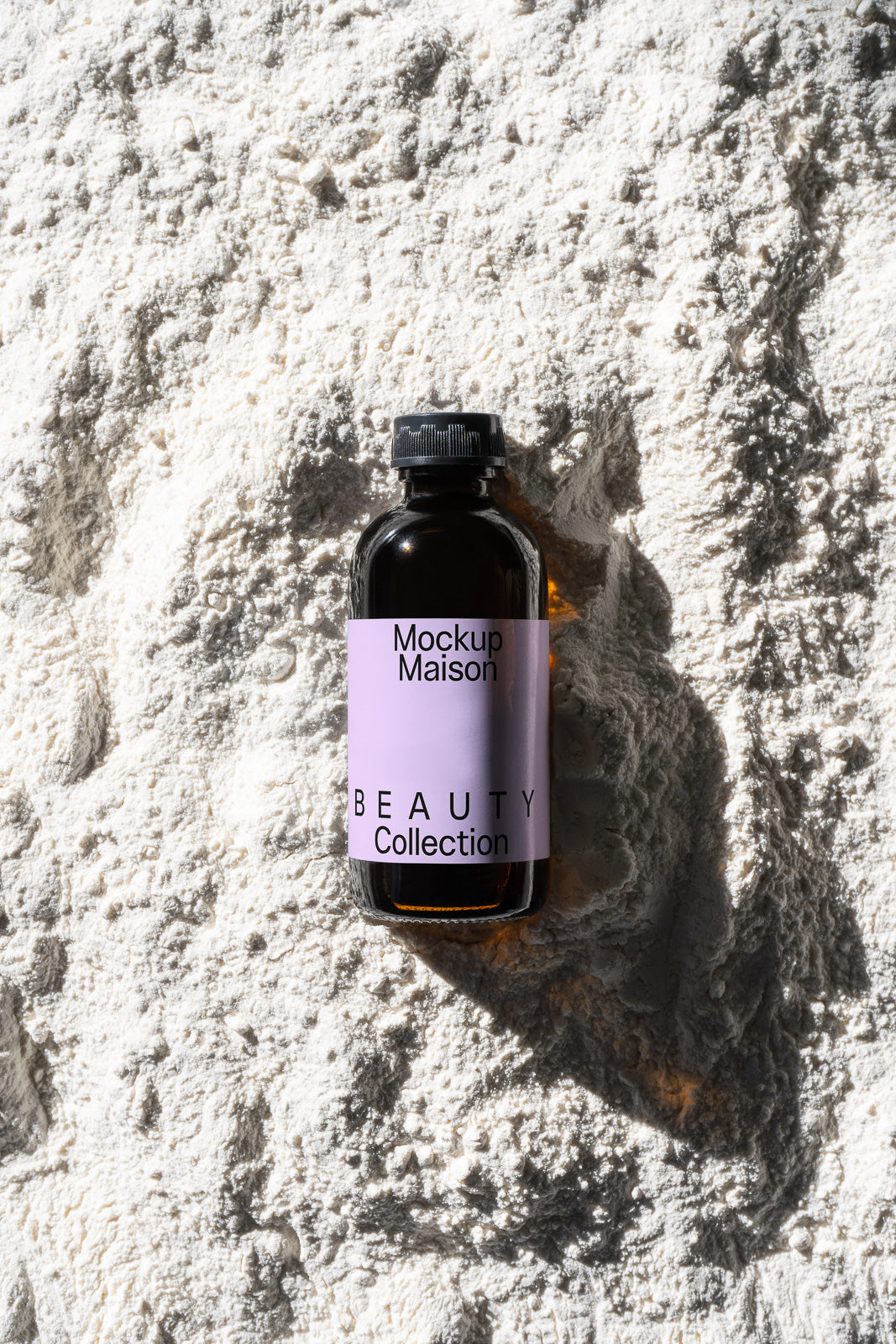 PSD mockup of a small amber bottle labeled "Mockup Maison Beauty Collection" resting on a textured white surface, casting a shadow.