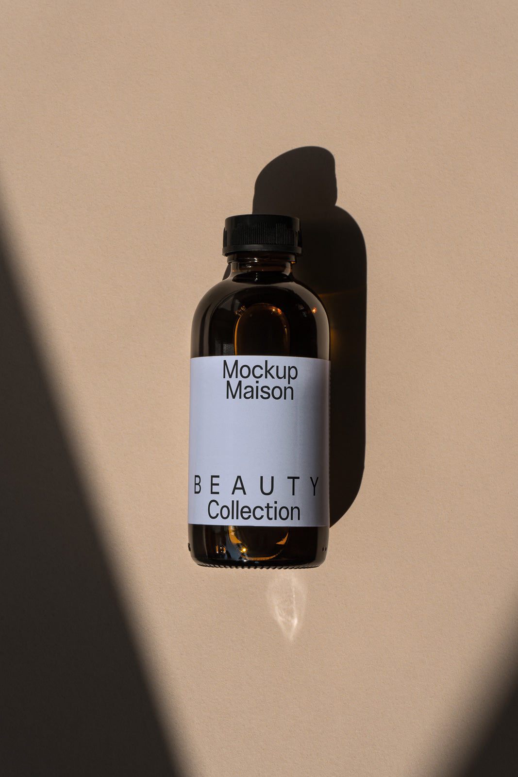 PSD mockup of an amber glass bottle with a black cap, labeled "Mockup Maison Beauty Collection," casting a shadow on a beige surface.