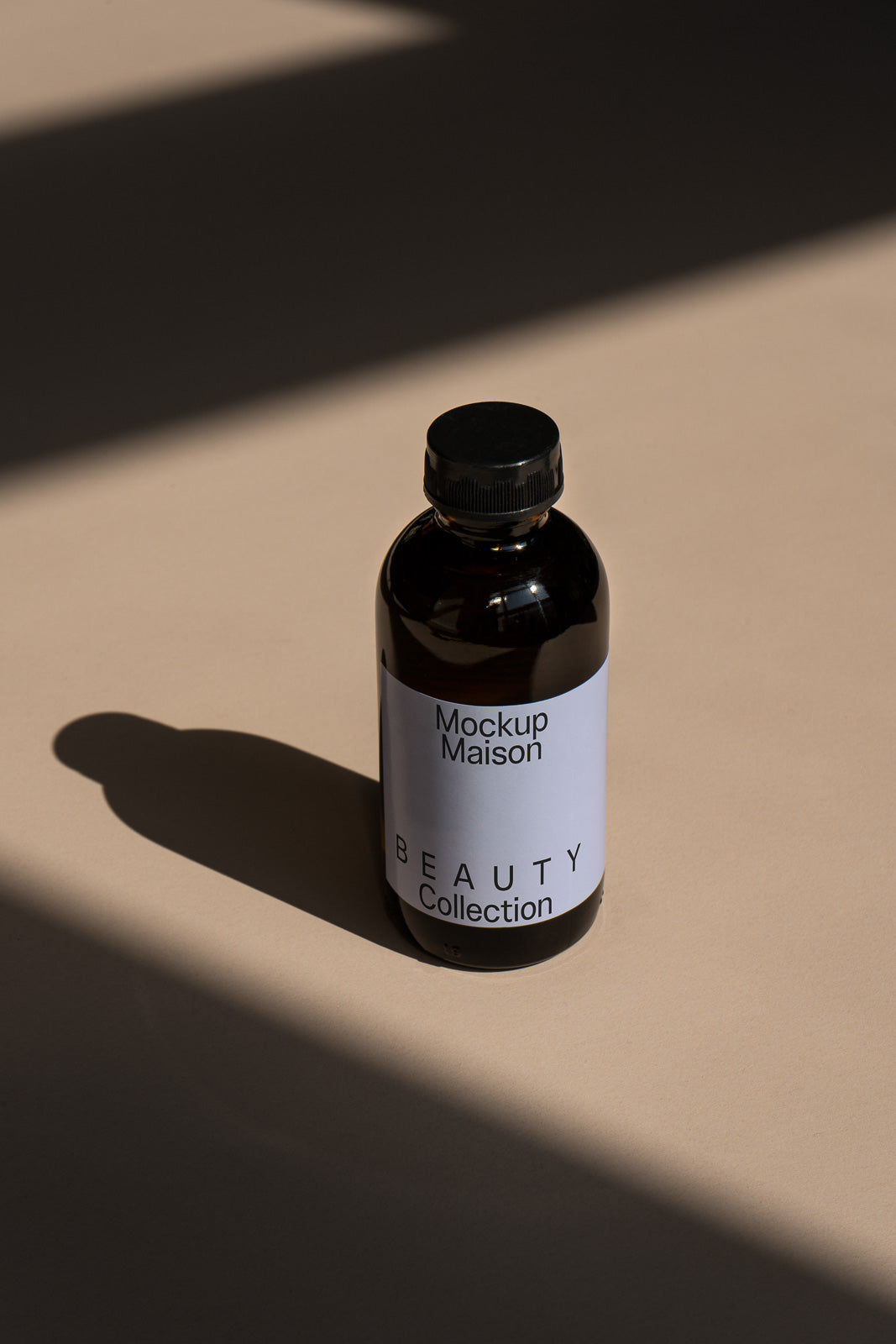 PSD mockup of a brown bottle with a black cap labeled "Mockup Maison Beauty Collection" standing on a beige surface with shadow patterns.