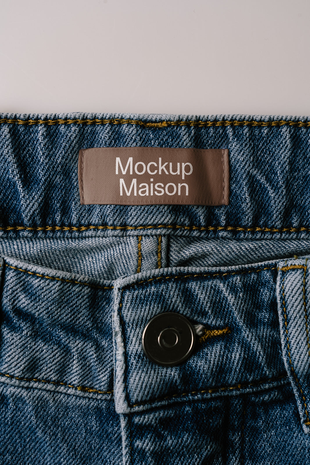 PSD mockup of a denim waistband close-up with a brown label reading "Mockup Maison" stitched above a button, showcasing fabric texture and detail.
