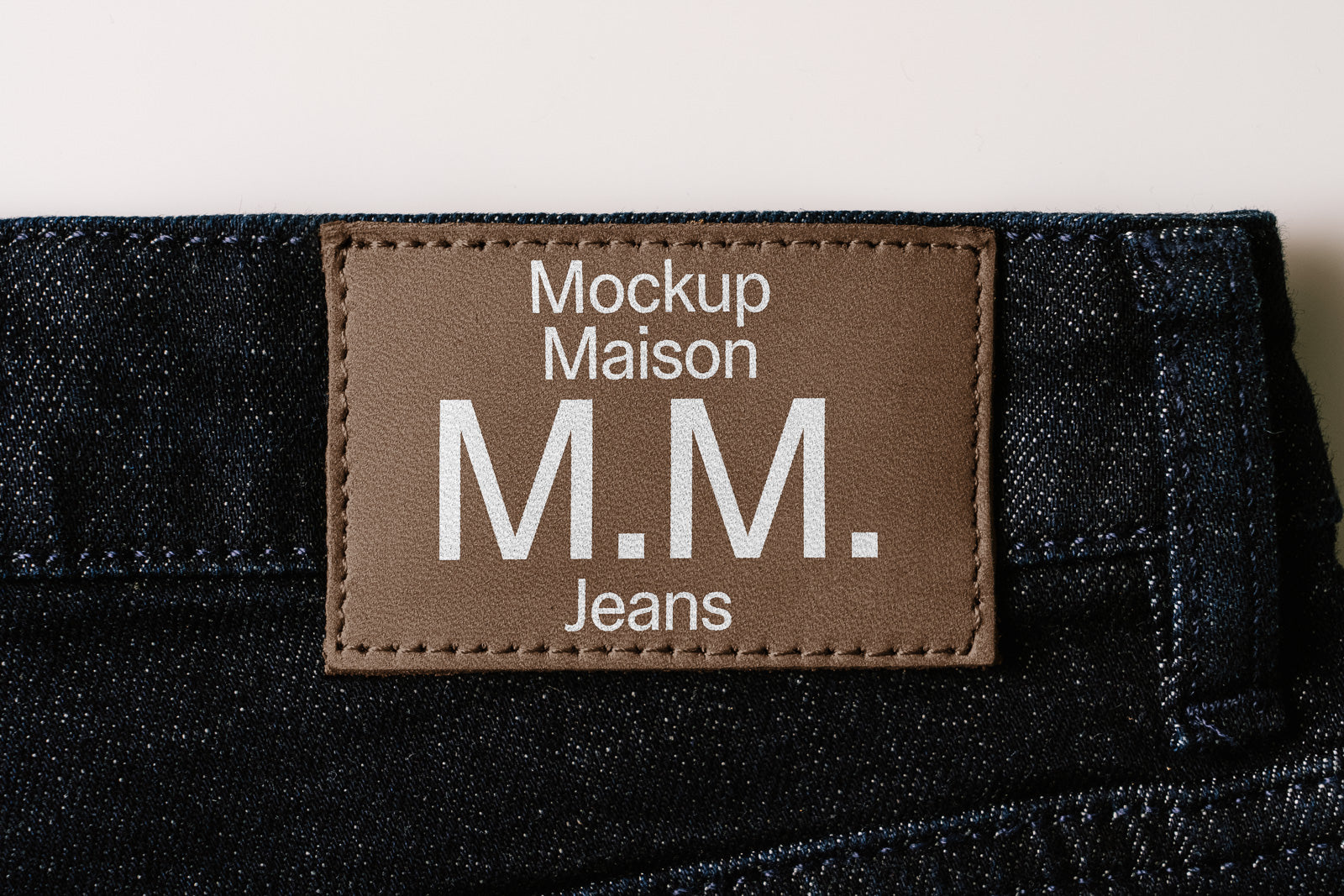 PSD mockup of a leather label on dark blue denim jeans, featuring the text "Mockup Maison M.M. Jeans" in white, stitched with dark thread.