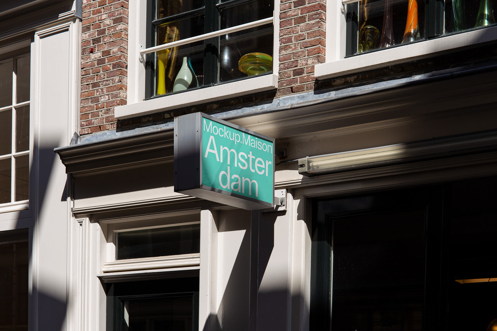 PSD mockup of a building facade with a turquoise rectangular sign displaying "Mockup Maison Amsterdam" in white text, surrounded by windows and brickwork.