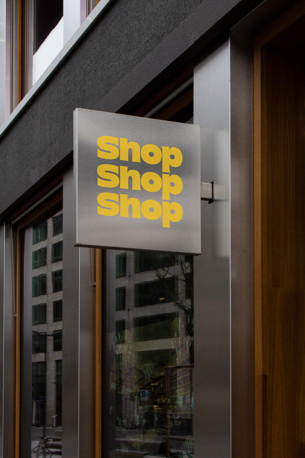 PSD mockup of a metal storefront sign with "Shop Shop Shop" in bold yellow letters, mounted on a modern building with glass windows.